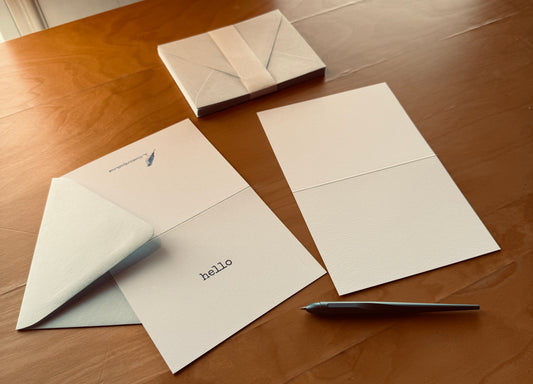 Open Bluebird Post “hello” card with blank interior, pastel blue envelopes, and pen on a wooden table.